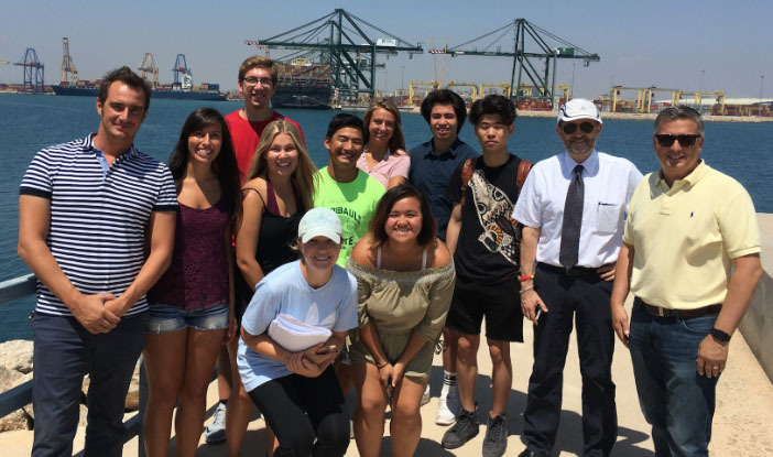 Students stand by a harbor with large cranes in the background.