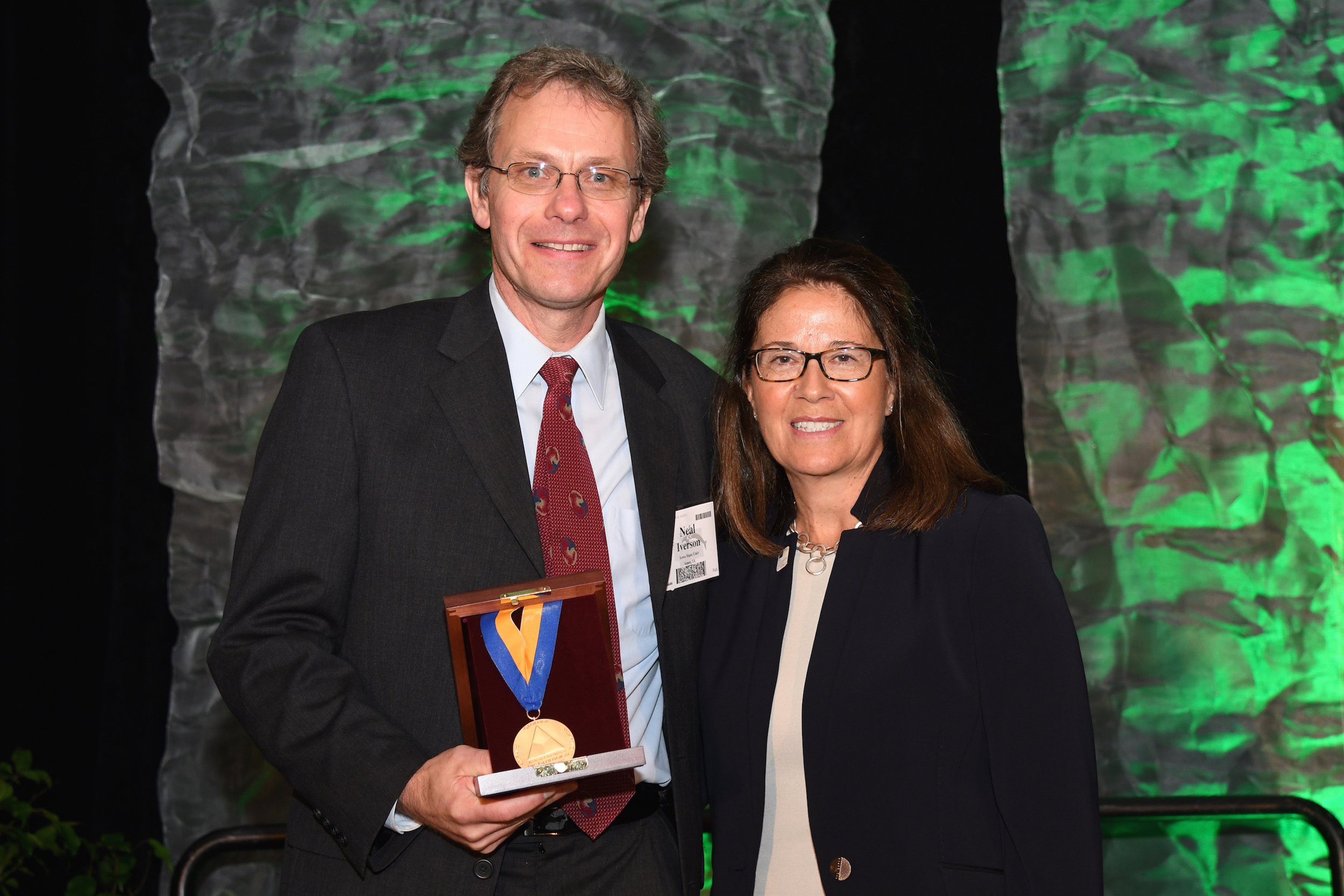 Neal Iverson holding a medal standing next to a woman in front of a green background.