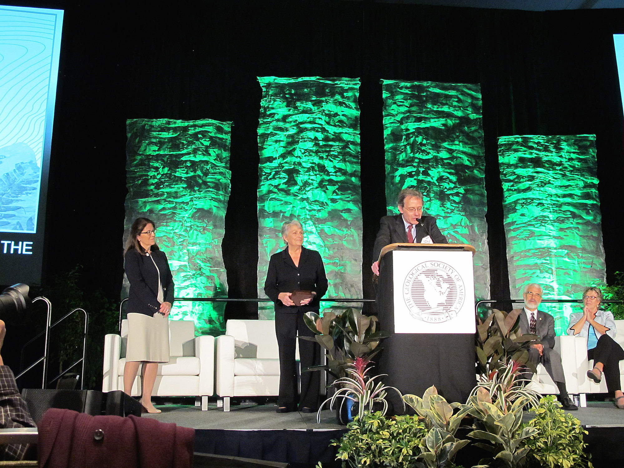 Neal Iverson stands behind a podium and plants on stage. A few people stand or sit in the back of the stage. The backdrop has four green column banners with rock patterns.