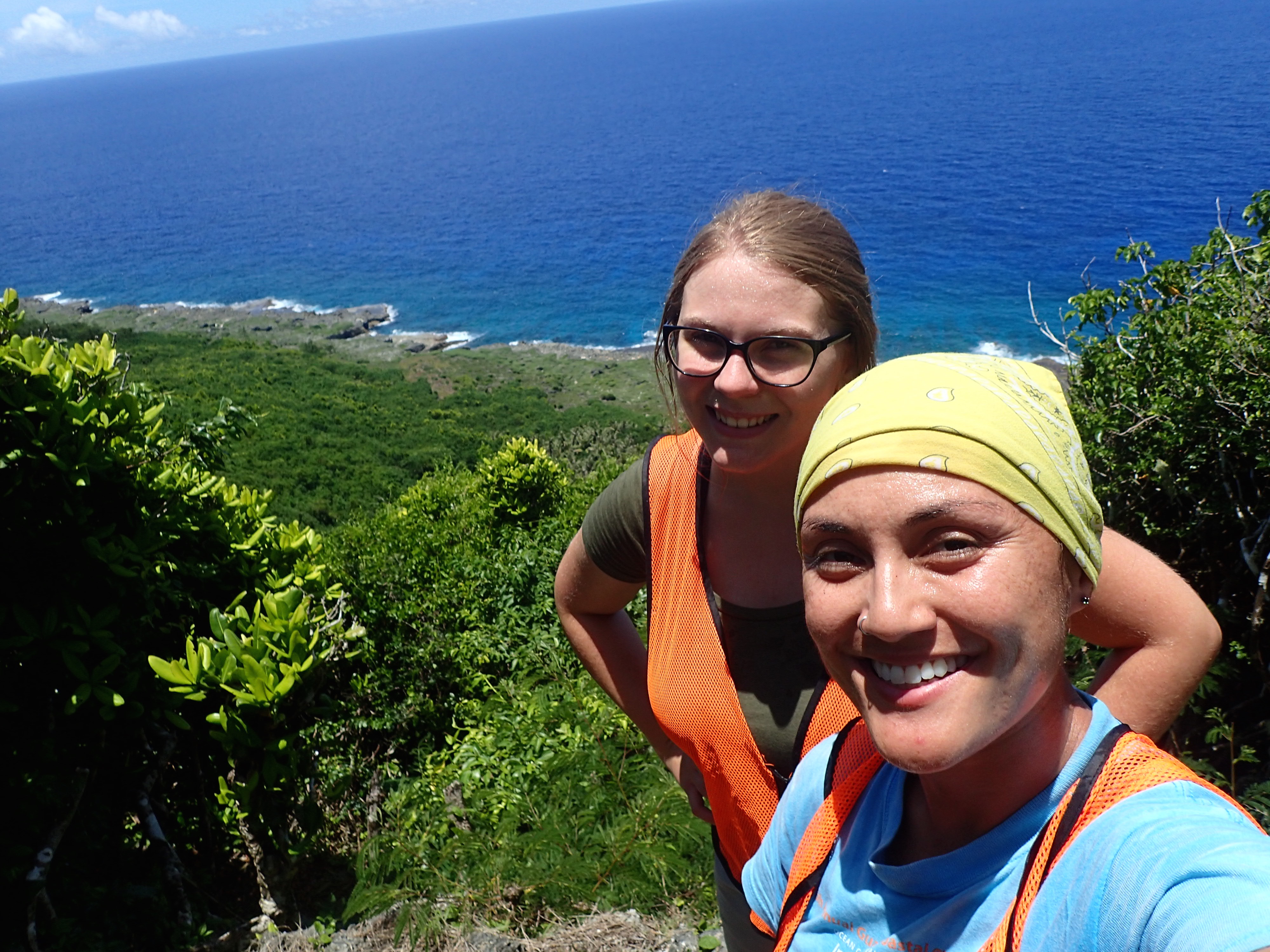 Two women stand in front of a cliff full of green vegetation, below and behind you can see the blue ocean.