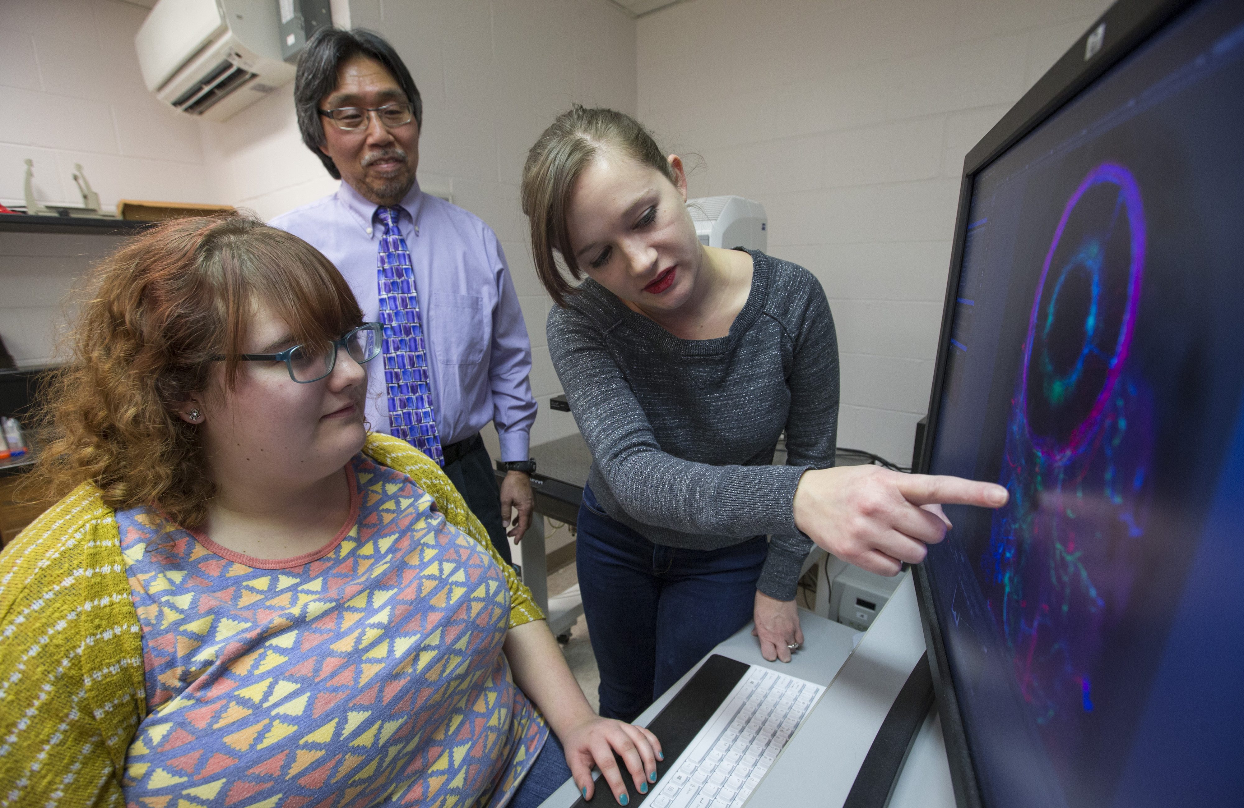Two students and a professor are gathered around a computer with a colorful display of an enlarged eye, one student points to the screen.