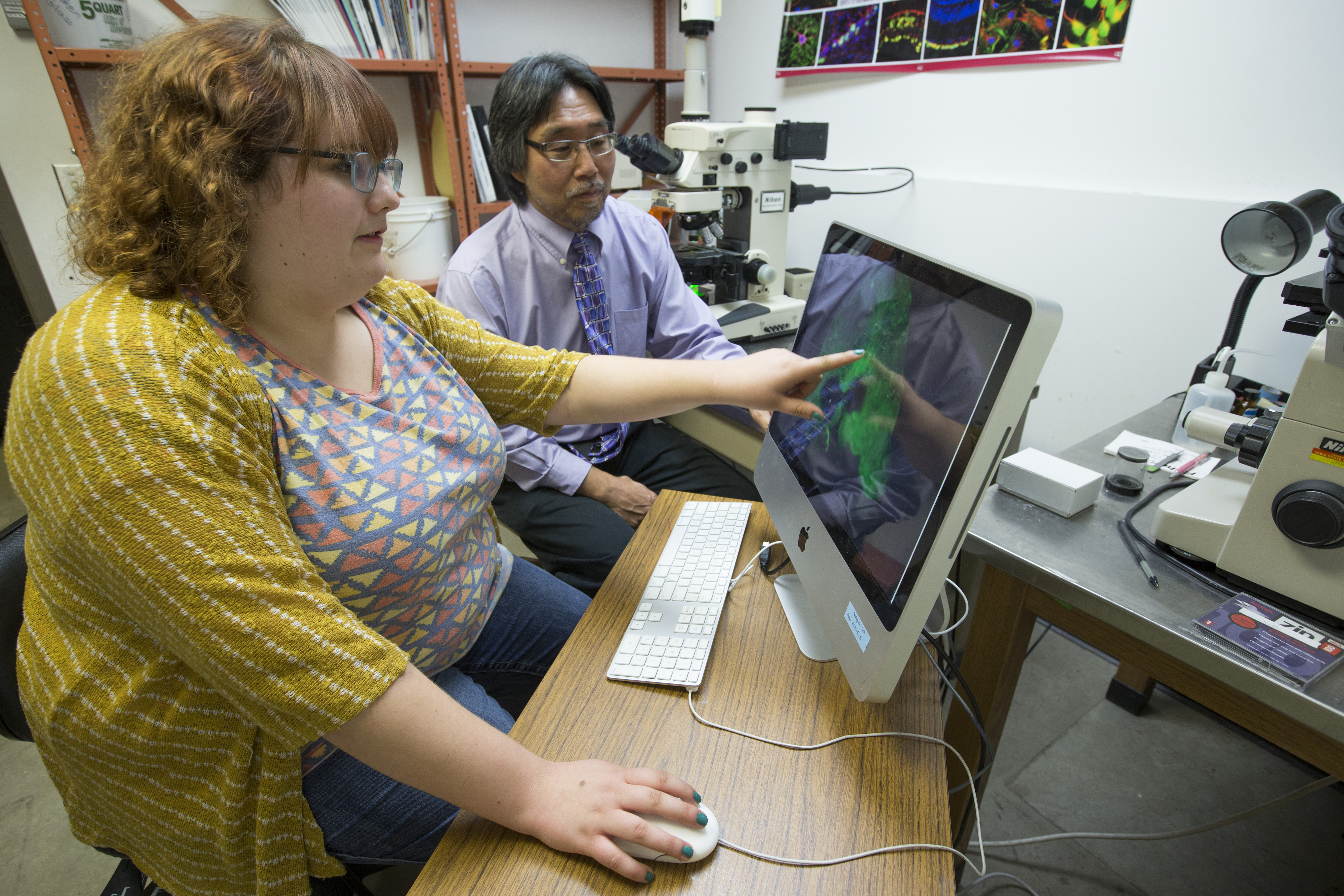 Kaelee Plante points at a computer image while Don Sakaguchi looks on. In the background are microscopes.