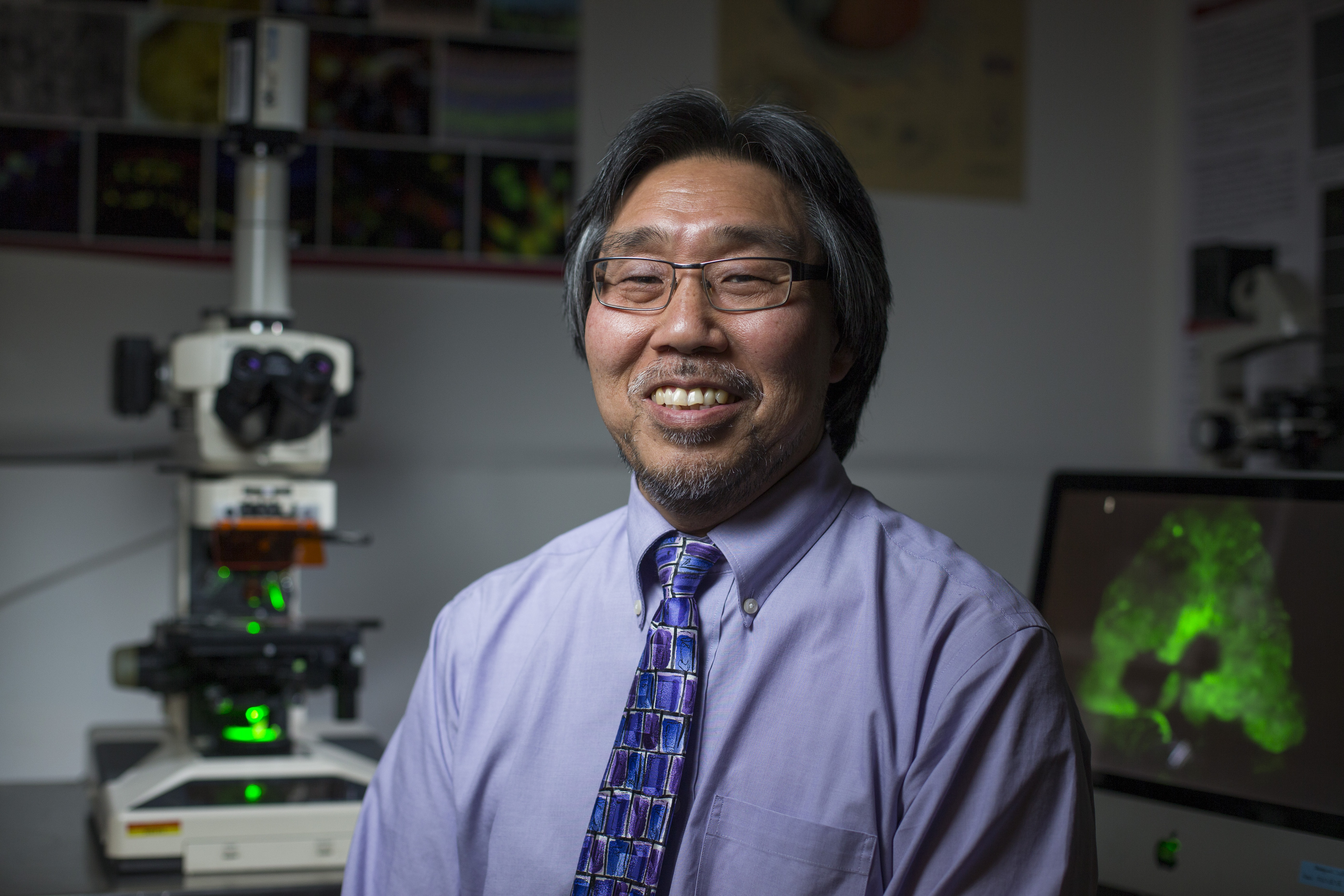 Don Sakaguchi with a microscope and computer imaging behind him.