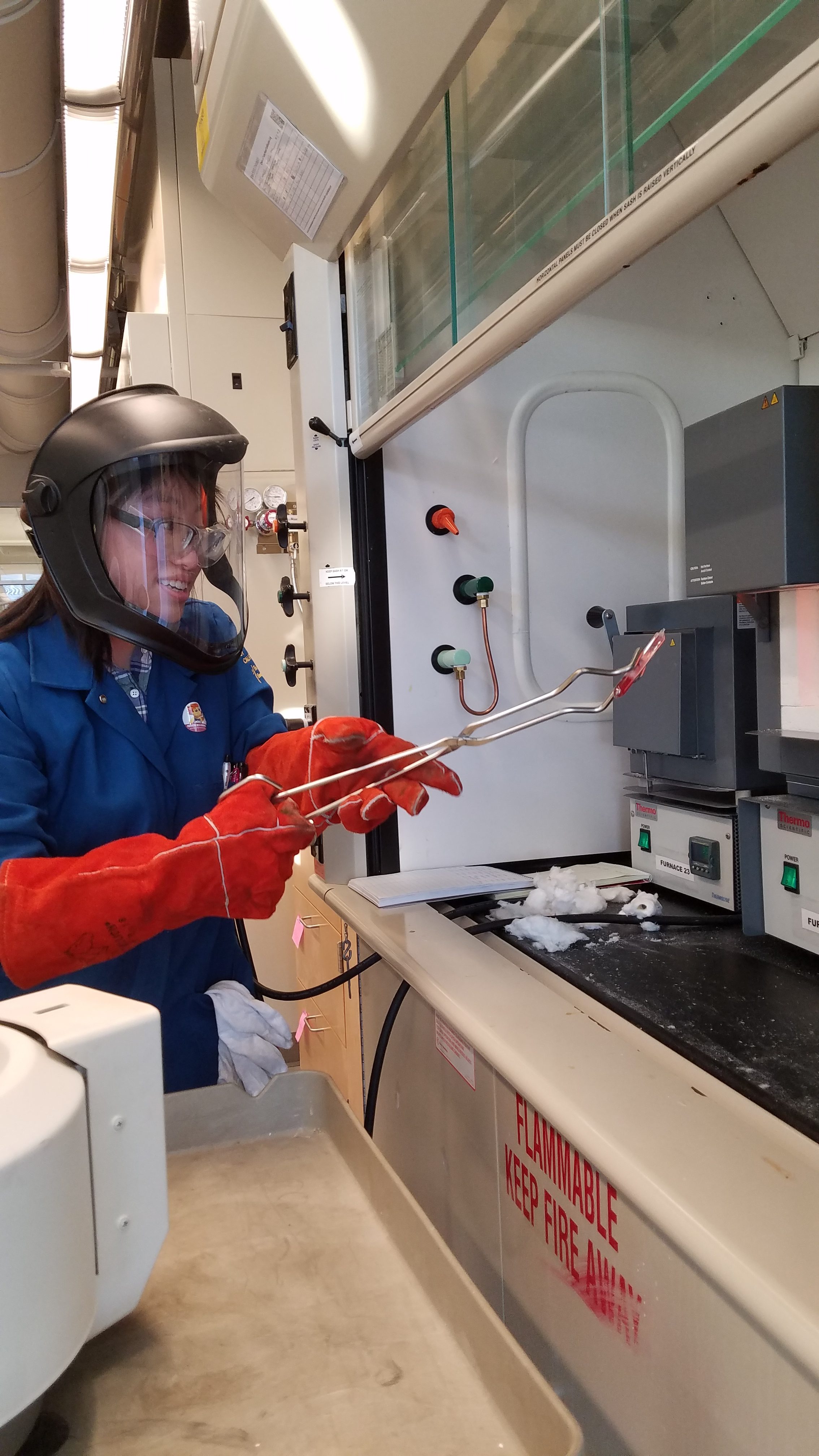 Katherine wears temperature protective gear and handles an item under the fumehood with tongs.