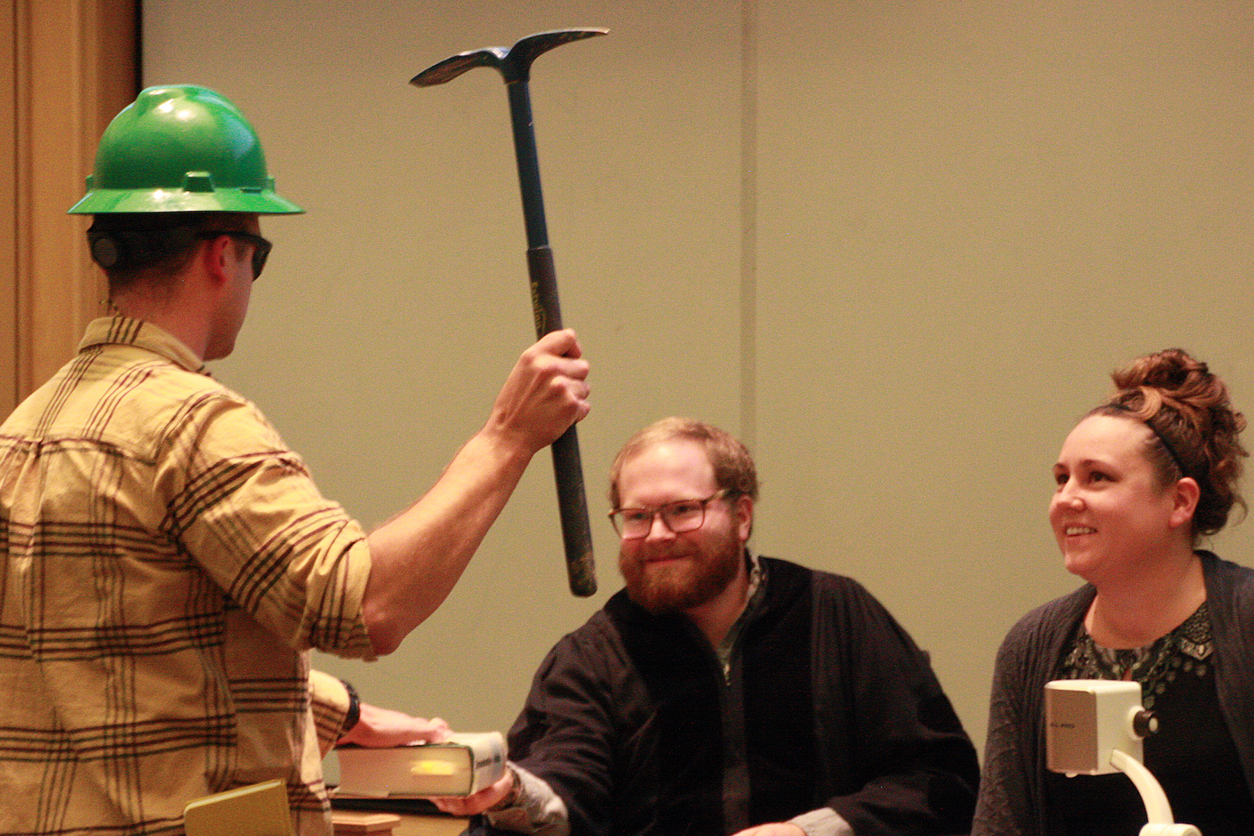 A man wearing a hard hat has his hand on a book and holds a pick-axe while two others look on.