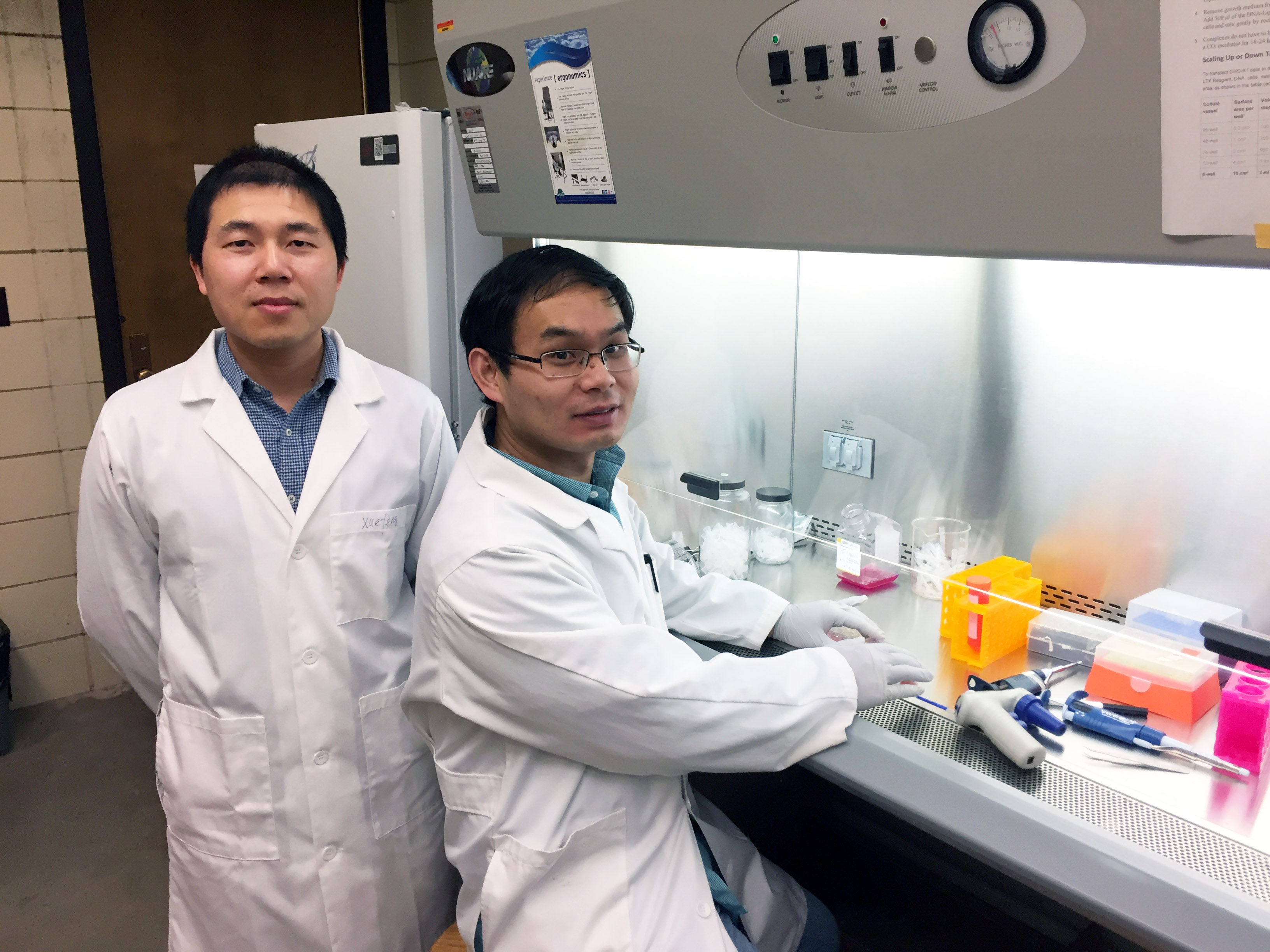 Two people are next to a fumehood in lab coats.