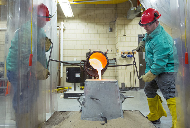 Two men in protective gear pour molten metal into a cast.