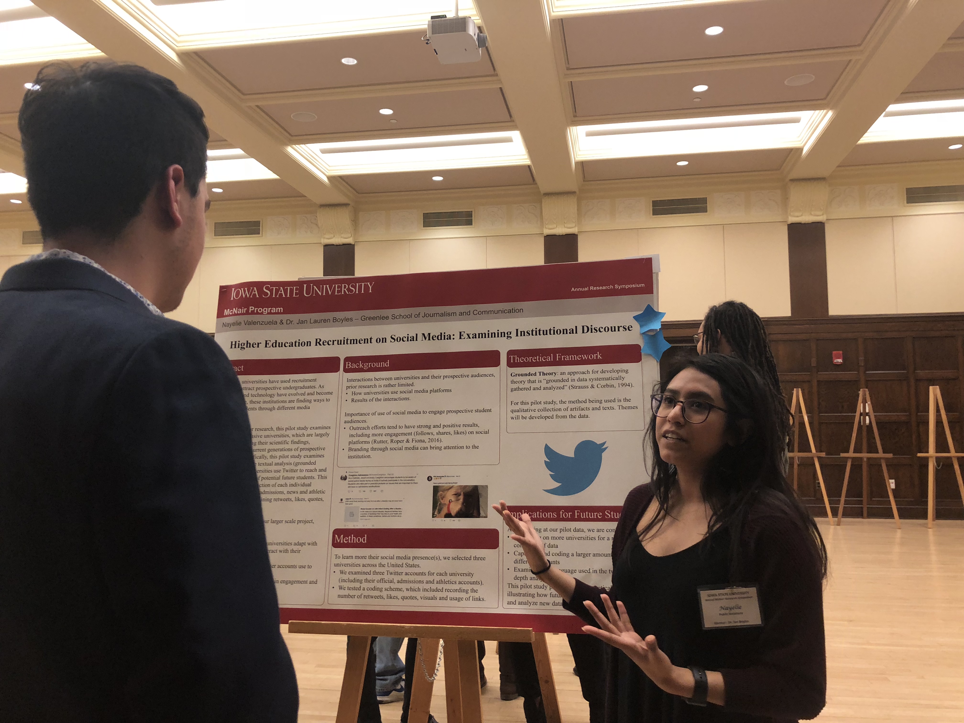 A student stands at a research poster explaining it to an onlooker.