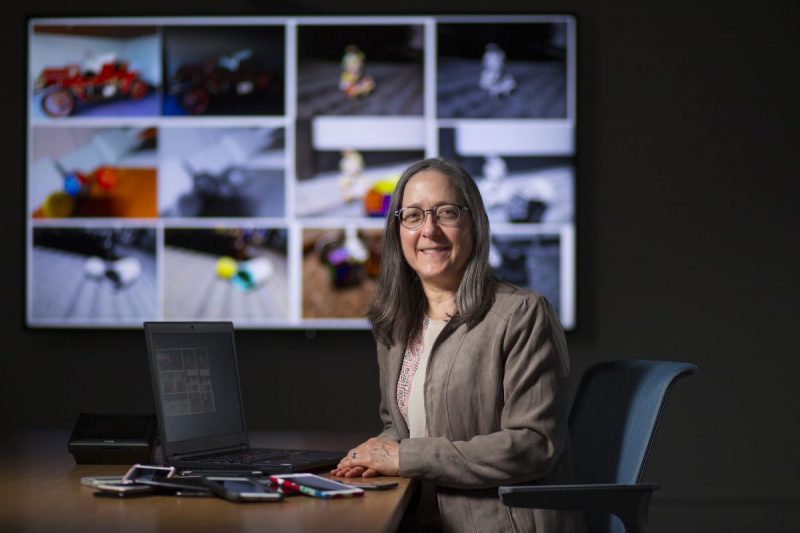 Jennifer Newman sits at a table with her laptop and a pile of phones. Behind her a screen shows images.