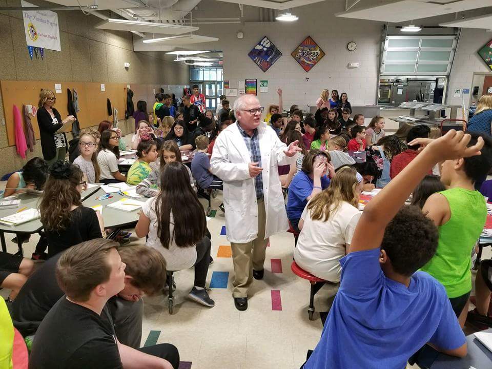 Joe Burnett in a lab coat stands mid students at tables.