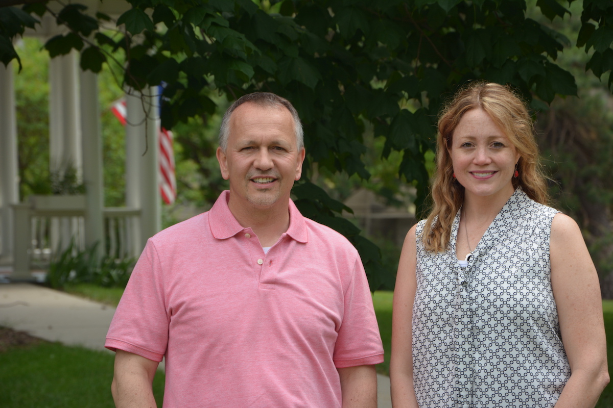 A man and woman stand outside. Trees, column, and a flag are in the background.