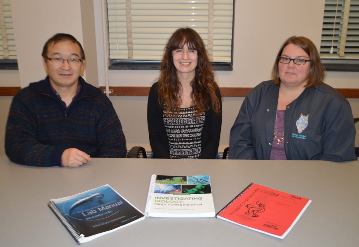 Three people sit at a table with three lab manuals in front of them.