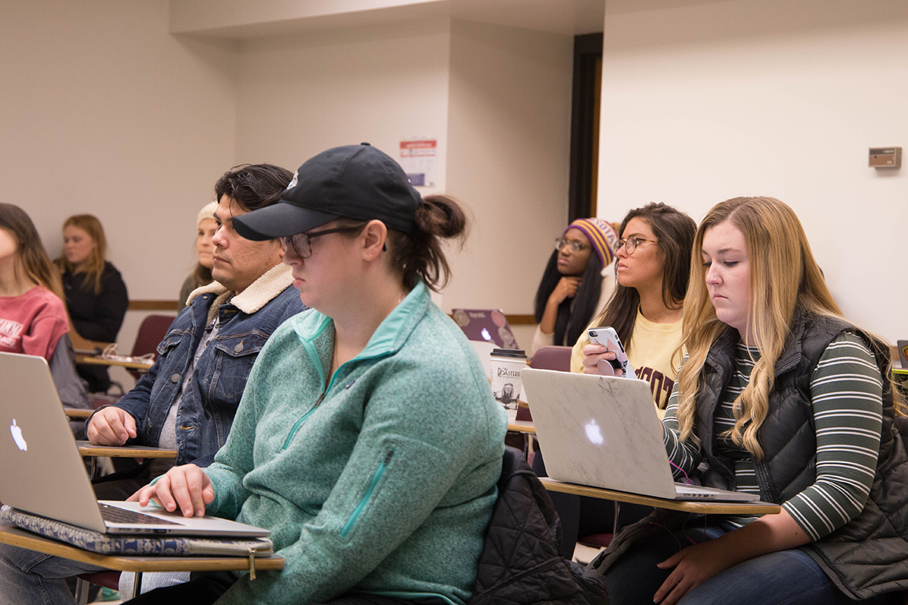 students sit at desks in class with computers