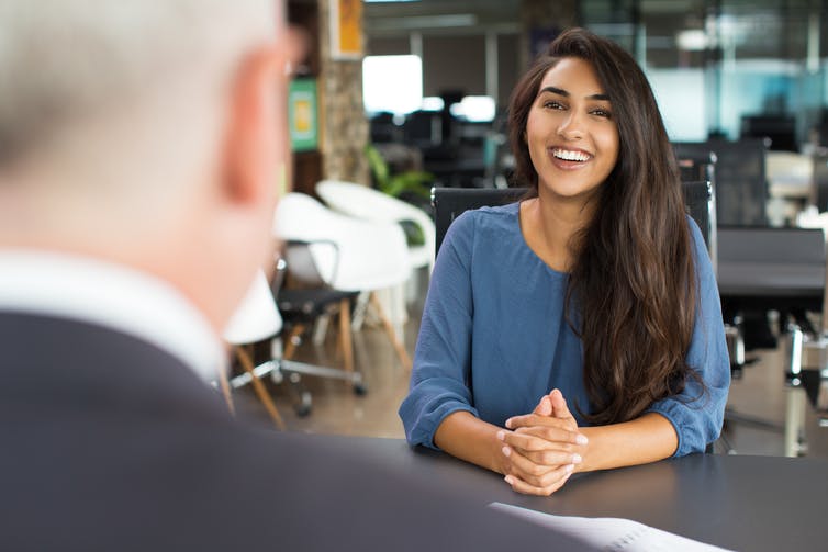 Photo of multicultural young woman talking across a desk. Photo shows partial back of head of man sitting across from her.