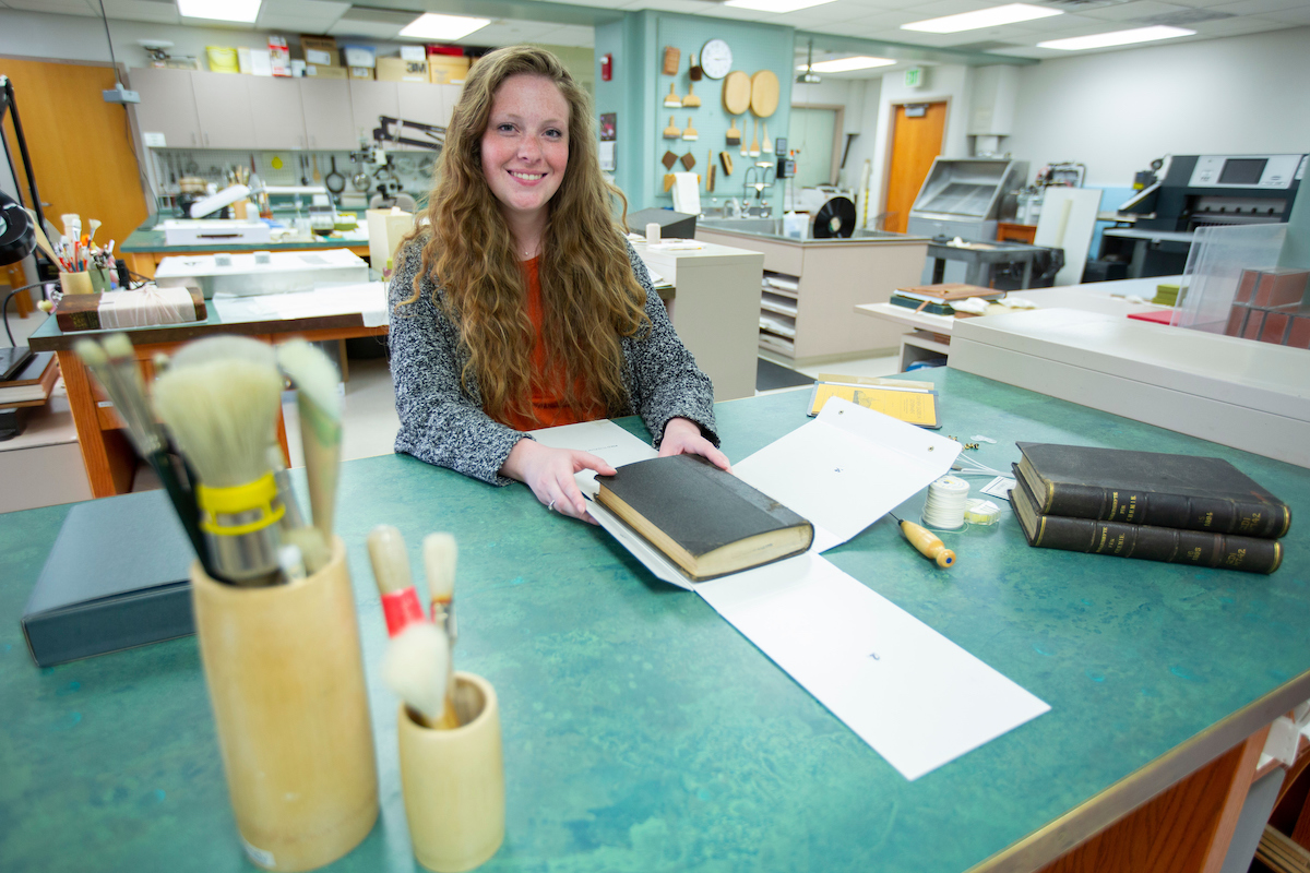 White woman with long hair working on a book in the library.