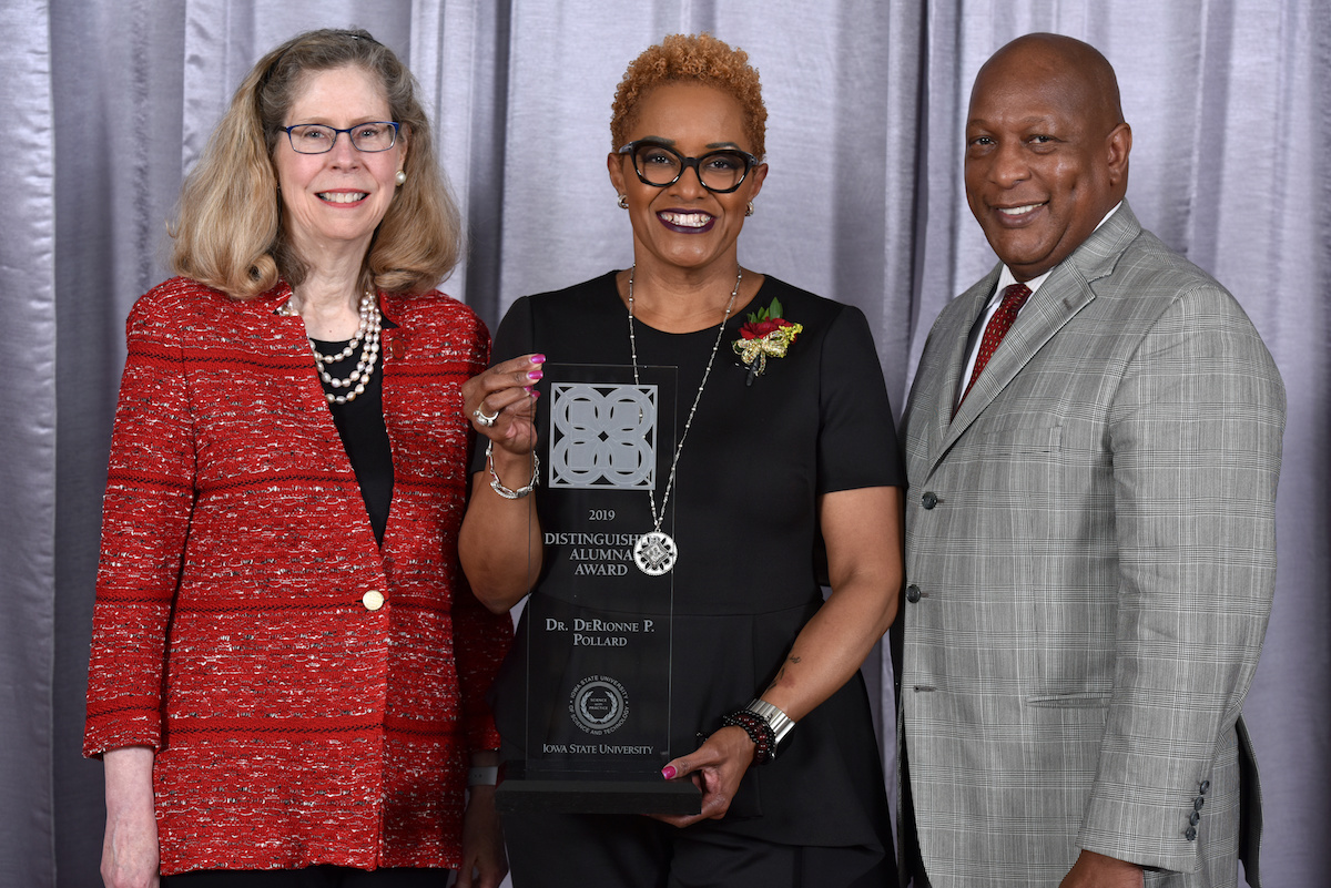 Three people, a while woman with blonde hair, a black woman with short blonde hair and a black man stand smiling at the camera. The woman in the middle holds an award.