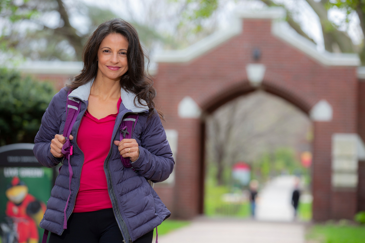 Woman with long brown hair wearing a backpack and smiling at the camera