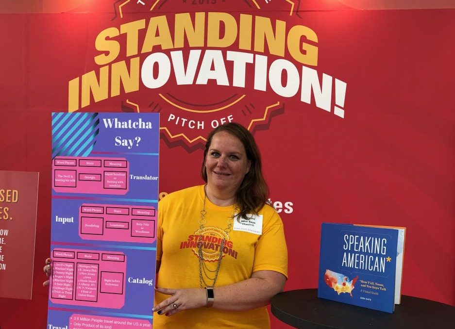 Student holding up display sign with an explanation of an app idea, in front of ISU Standing Innovation backdrop, next to table with book