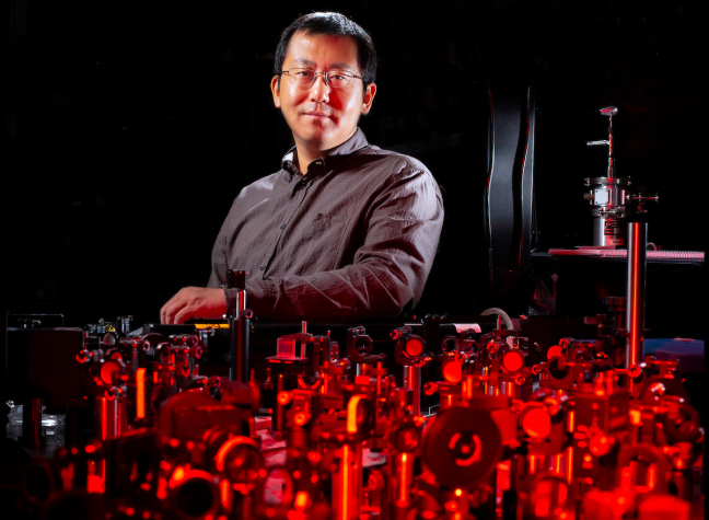 Jigang Wang standing behind lasers and other light measuring equipment in his laboratory.