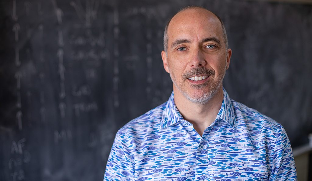 Robert McQueeney stands in front of a classroom blackboard