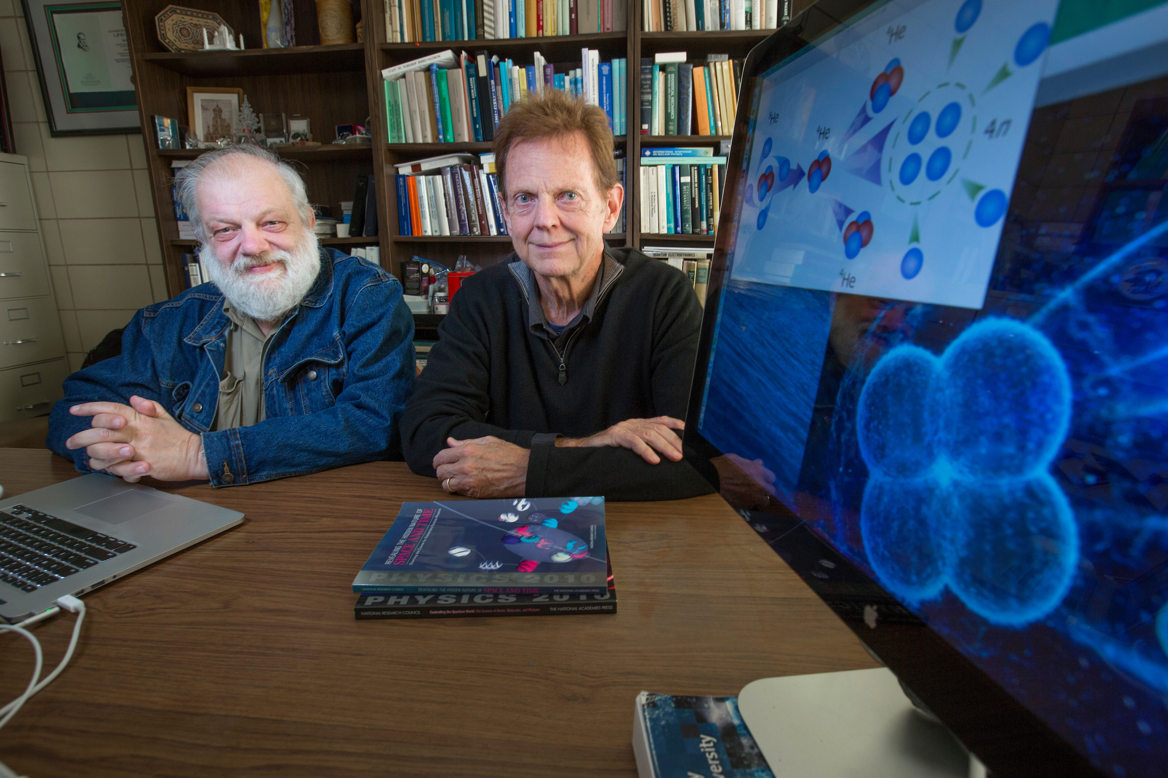 Scientists James Vary and Andrey Shirokov pose in front of a computer image of a four-neutron structure.