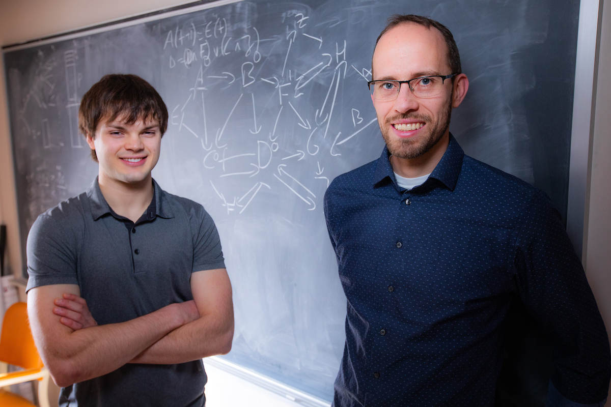 Claus Kadelka and an undergraduate research student stand in front of blackboard in a classroom.