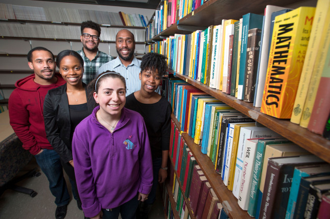 Mathematics graduate students and MOCA members (counter-clockwise from top) Michael Dairyko, Derek Young, Chassidy Bozeman, Erica Johnson, Shanise Walker and assistant professor Michael Young in the mathematics reading room standing by a wall filled with shelves of books.