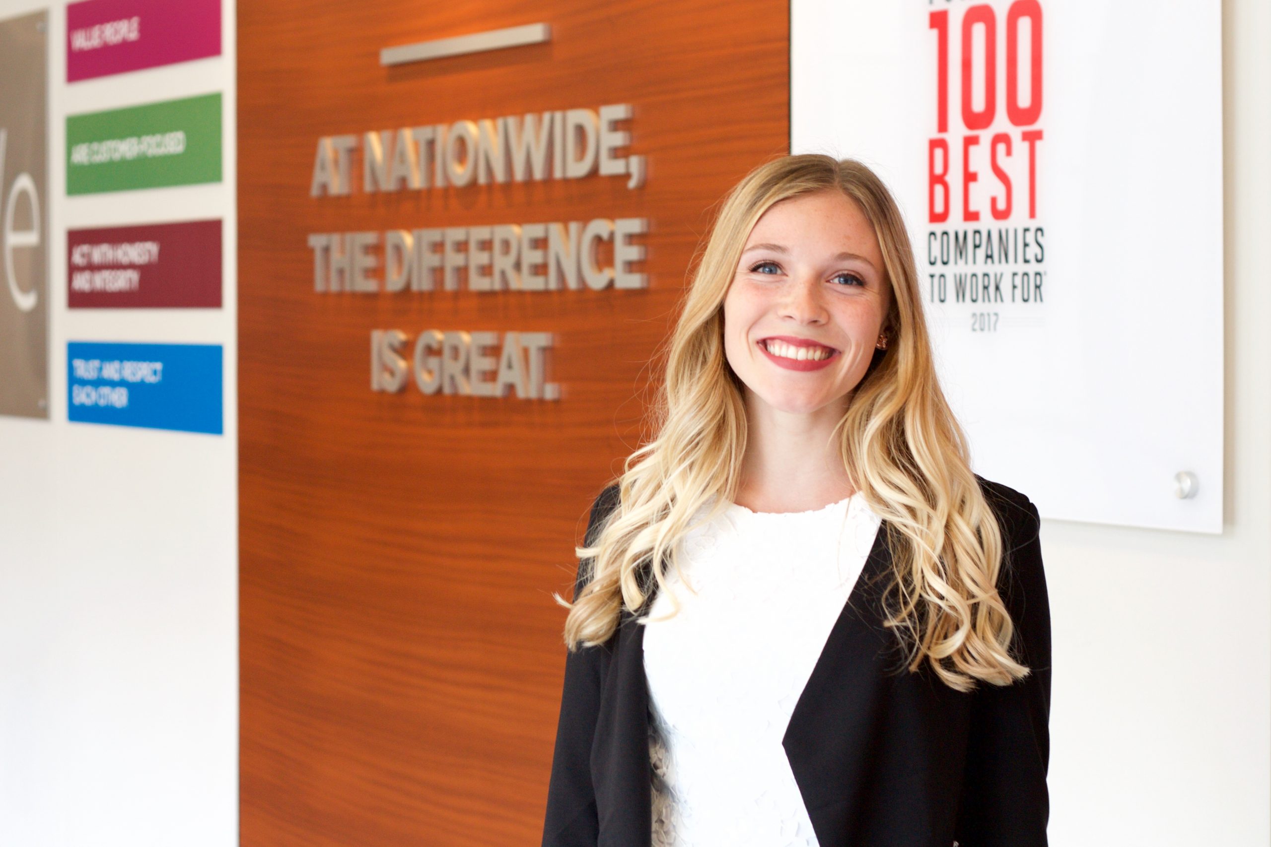 Undergraduate student Aimee Rodin poses at her internship at Nationwide Insurance Company. On the wall behind her the company has a sign that it is part of 100 Best Companies to Work For.
