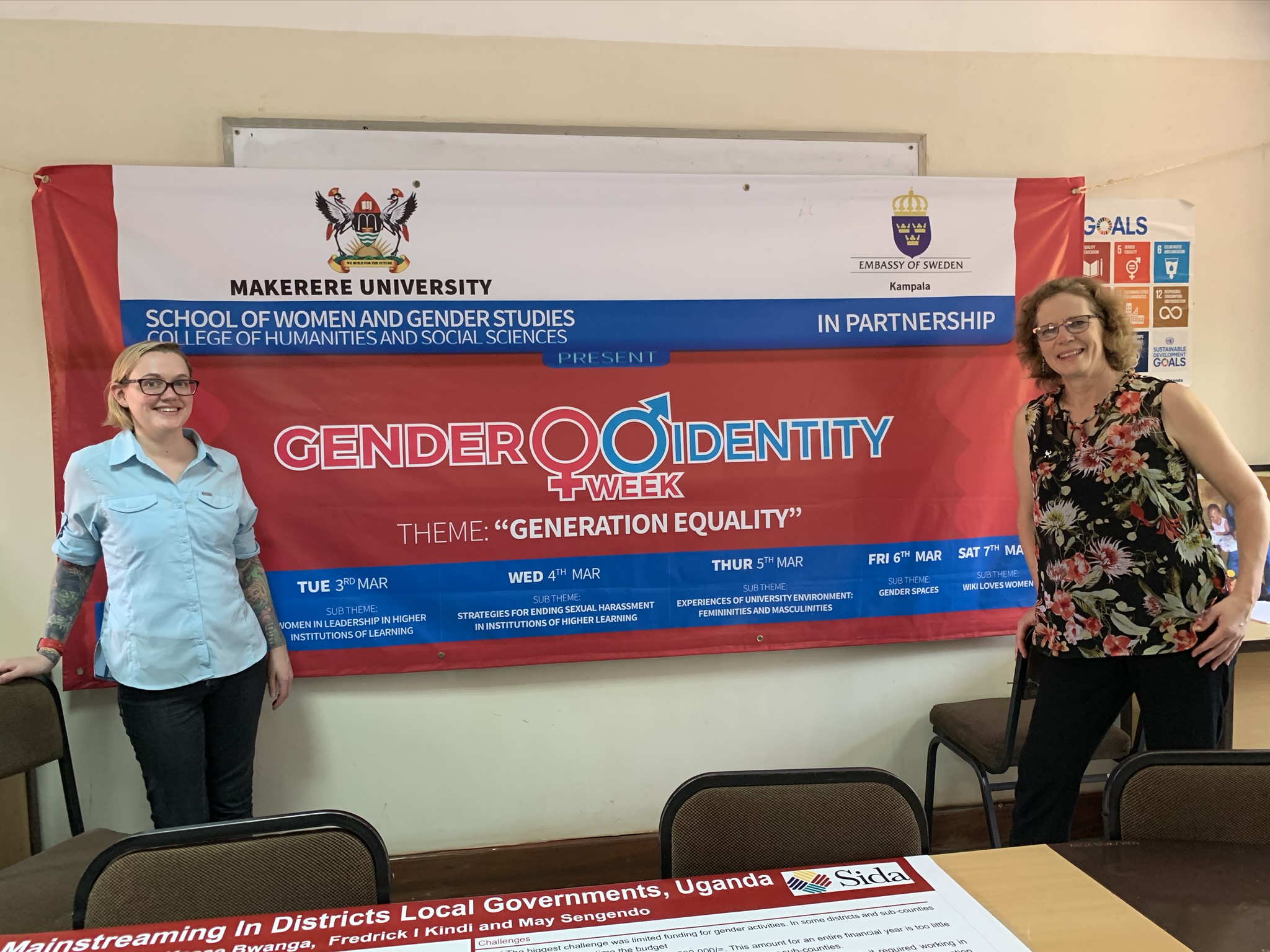 two white women smiling, standing in a classroom in front of a banner which reads "Gender Identity Week"